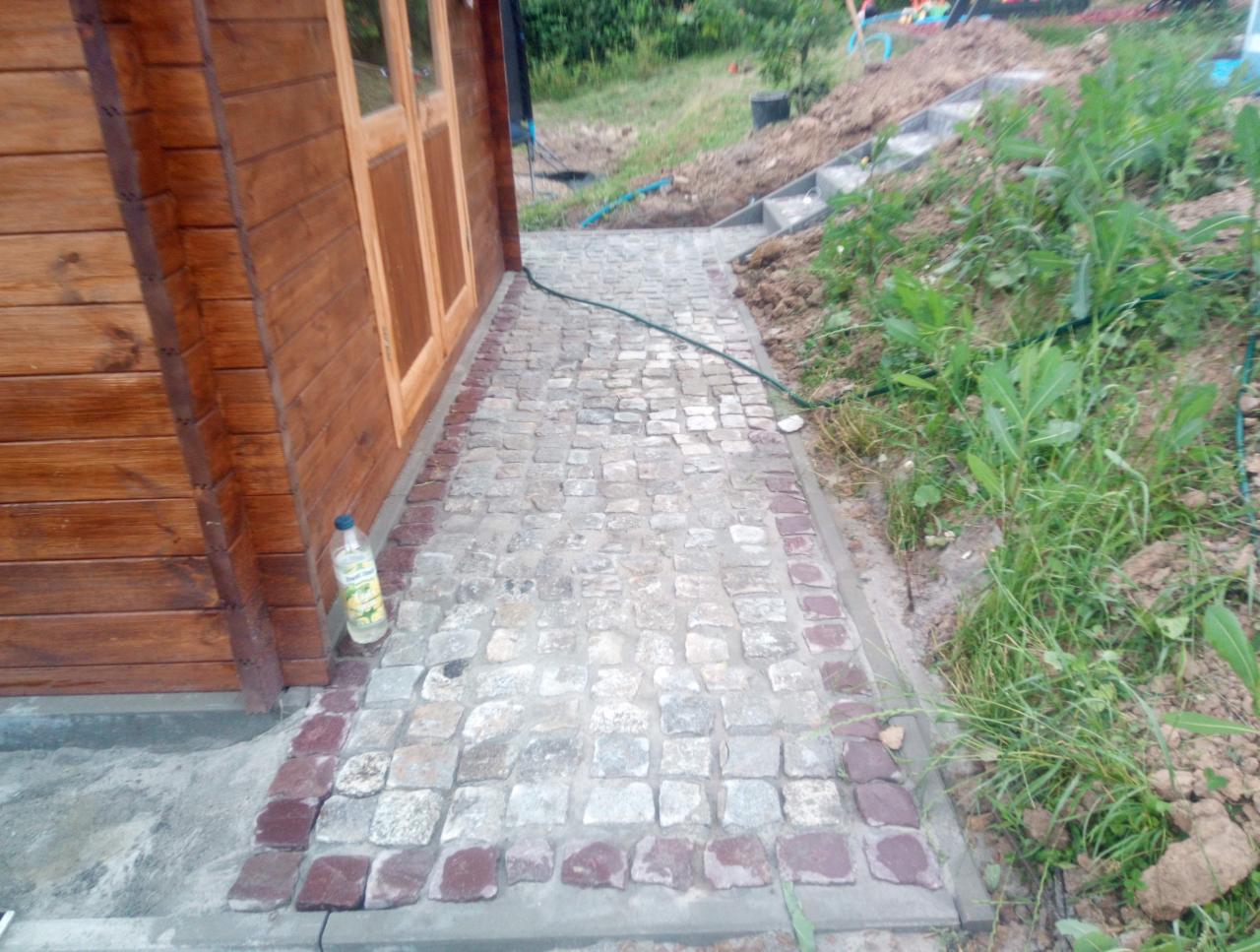 Ścieżka z kostki granitowej prowadząca do drewnianego domku, z obrzeżem z ciemniejszej kostki, widoczna butelka wody przy ścianie i schody na wzniesieniu w tle.
