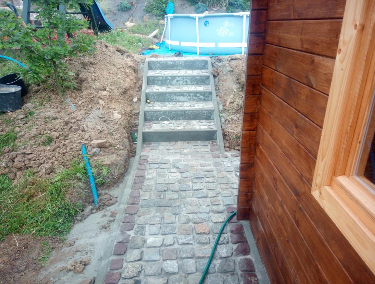 Ułożona ścieżka z kostki granitowej prowadząca do betonowych schodów, obok drewnianej konstrukcji i basenu w tle.