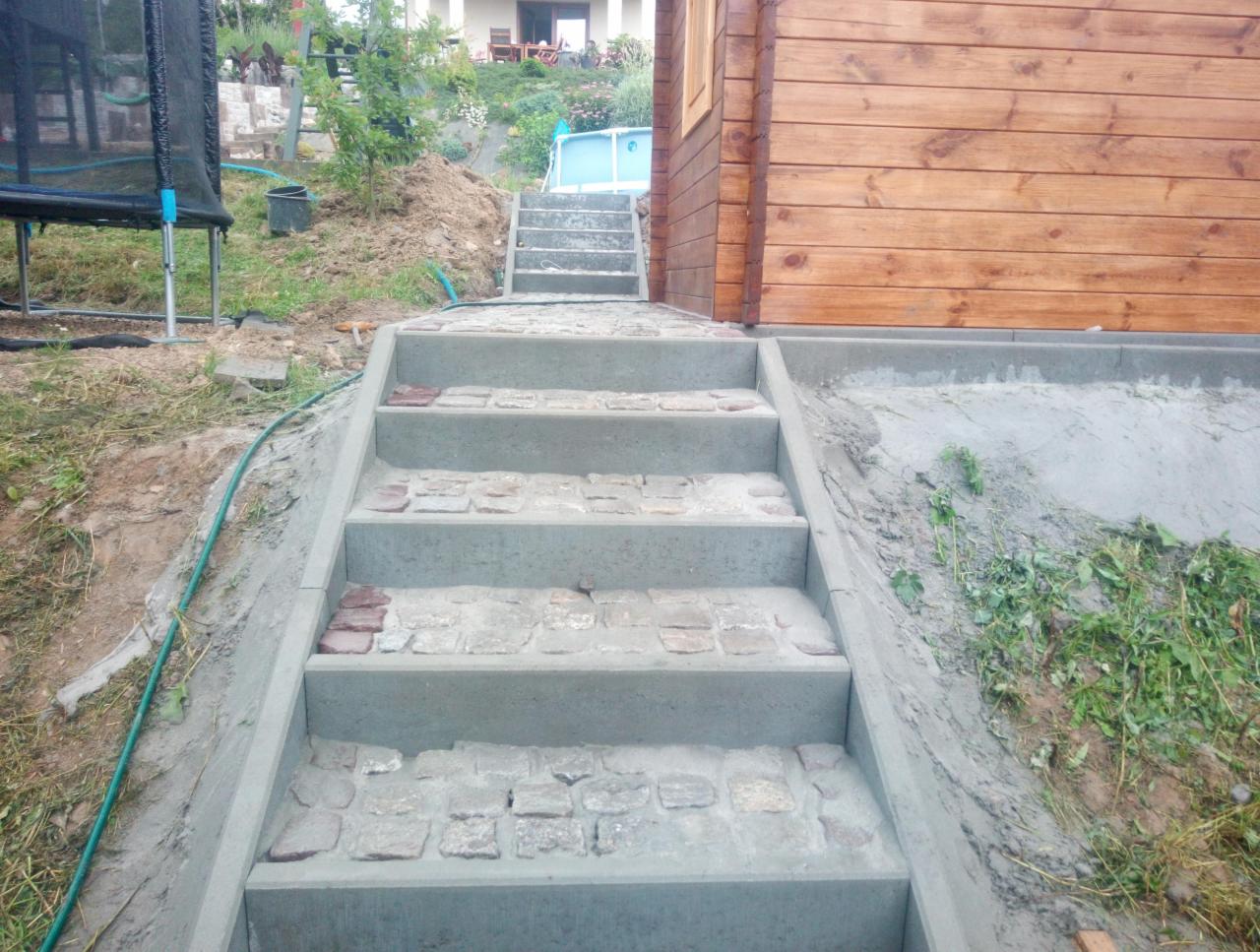 Betonowe schody z wstawkami z kostki granitowej prowadzące do drewnianego domku, widok z dołu, w tle trampolina i basen.