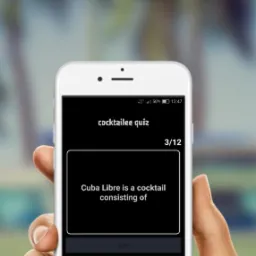 Smartfon w dłoni z uruchomionym quizem o nazwie 'Cocktailee Quiz', pytanie o skład drinka Cuba Libre, trzy opcje odpowiedzi: rum, wódka, whisky.