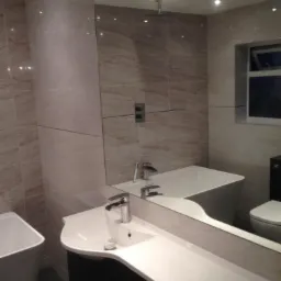 Wykończona łazienka z beżowymi płytkami imitującymi marmur, umywalka z czarną szafką, lustro odbijające wannę i toaletę.