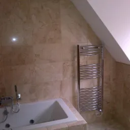 Łazienka na poddaszu z wanną obudowaną płytkami trawertynowymi, grzejnik drabinkowy na ścianie, beżowe płytki na ścianach i podłodze.