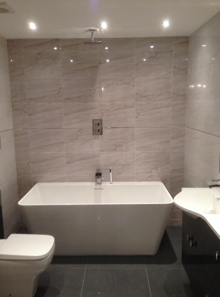 Nowoczesna łazienka z wolnostojącą białą wanną na tle beżowych płytek imitujących marmur, widoczna również umywalka z szafką w kolorze czarnym i toaleta.