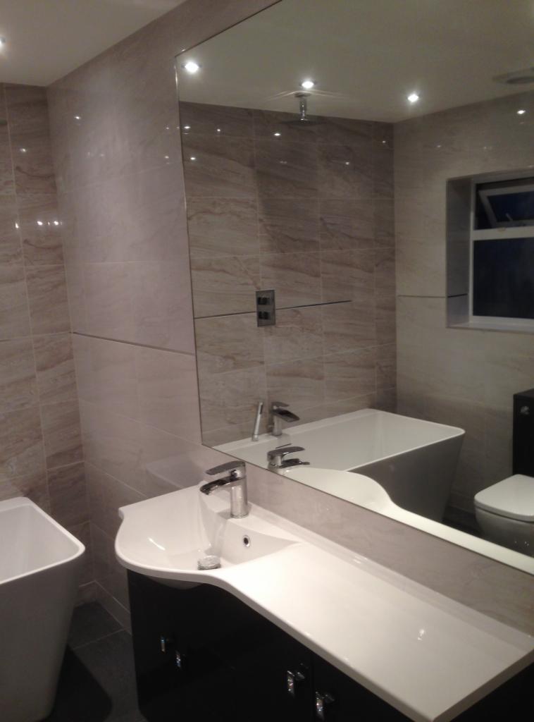 Wykończona łazienka z beżowymi płytkami imitującymi marmur, umywalka z czarną szafką, lustro odbijające wannę i toaletę.