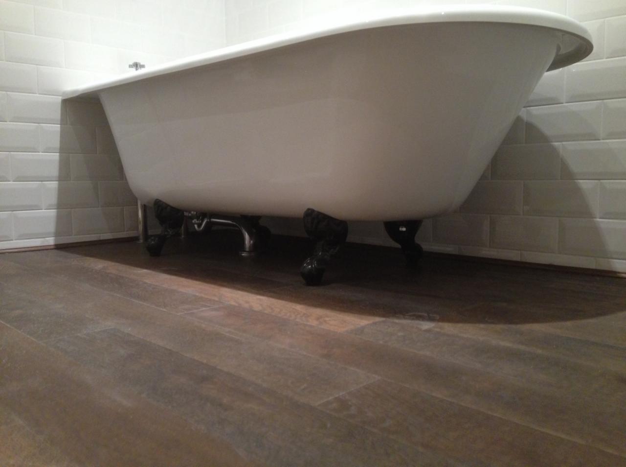 Biała wanna wolnostojąca na ozdobnych, czarnych nóżkach, z widoczną chromowaną armaturą, ustawiona na podłodze z paneli imitujących drewno, z białymi płytkami w tle.