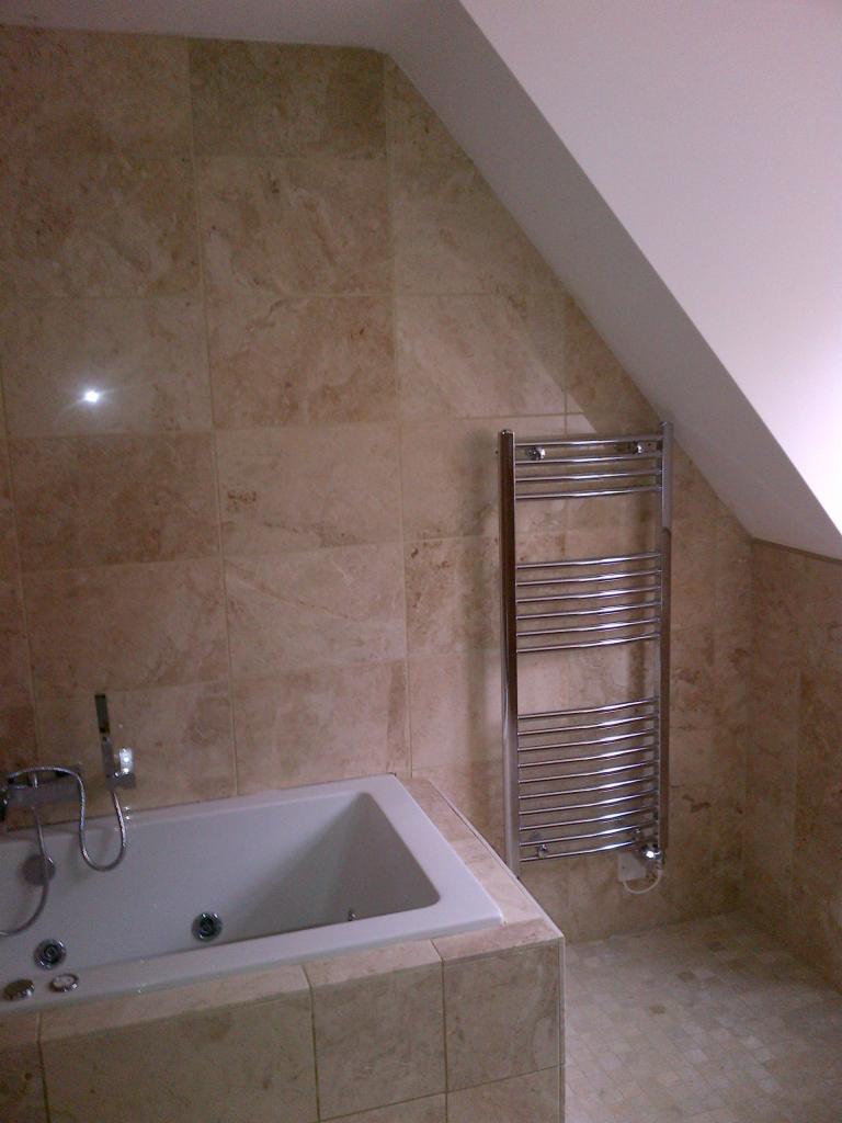 Łazienka na poddaszu z wanną obudowaną płytkami trawertynowymi, grzejnik drabinkowy na ścianie, beżowe płytki na ścianach i podłodze.