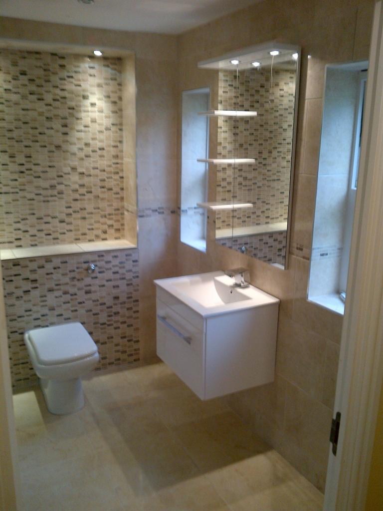 Jasna łazienka z beżowymi płytkami mozaikowymi, podwieszana szafka z umywalką, toaleta oraz lustro z półkami i oświetleniem LED.