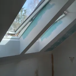 Montaż okien dachowych w skośnym suficie, widoczne szpachlowane połączenia, jasne wnętrze z widokiem na drzewa przez okno.