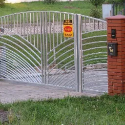 Metalowa brama wjazdowa z dekoracyjnym wypełnieniem w kształcie wachlarza, osadzona na ceglanych słupkach z zamontowaną skrzynką na listy i tabliczką ostrzegawczą.