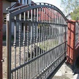 Metalowa brama wjazdowa z ozdobnymi elementami, zamontowana na murowanych słupkach z czerwonej cegły, widok z boku na tle zabudowań i zieleni.