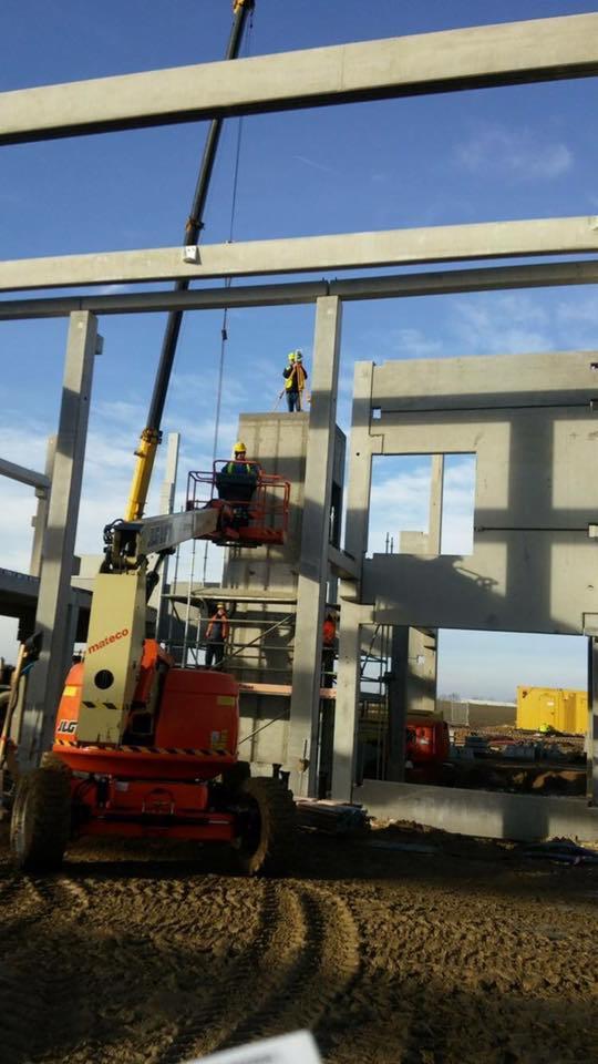 Montaż prefabrykowanych elementów betonowych szkieletu budynku z użyciem dźwigu i podnośnika koszowego; pracownicy w kaskach i kamizelkach odblaskowych.
