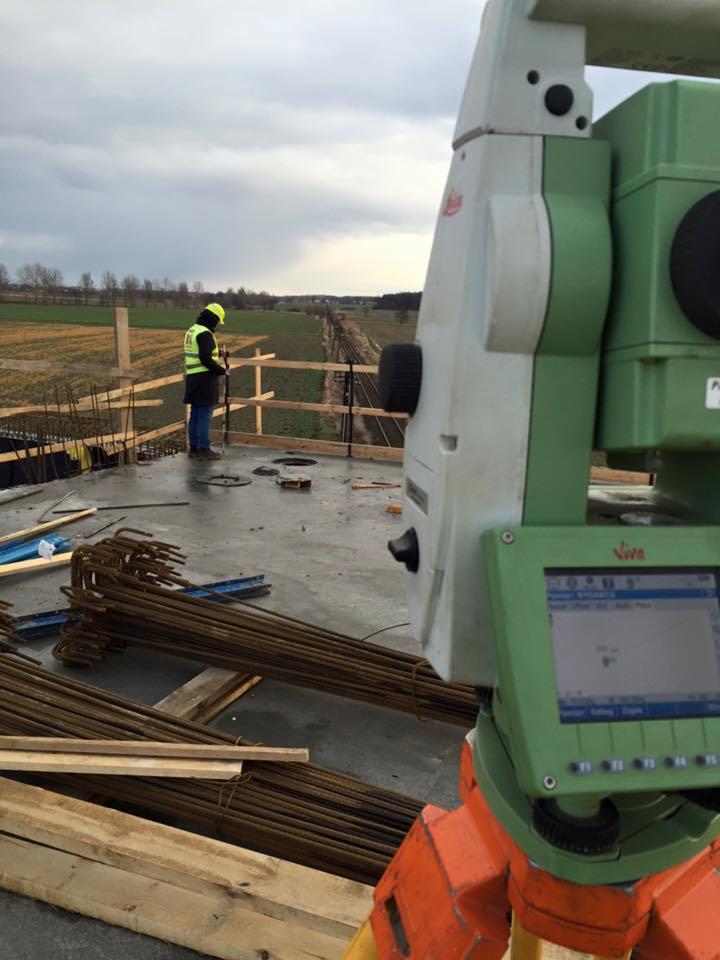 Pomiar geodezyjny na budowie: teodolit Leica Viva na pierwszym planie, w tle pracownik w kamizelce odblaskowej wykonujący pomiary na betonowej płycie z wystającymi prętami zbrojeniowymi.