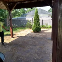 Częściowo ułożona kostka brukowa pod drewnianą altaną, zasypana piaskiem, z widocznym fragmentem ogrodu i tujami w tle.