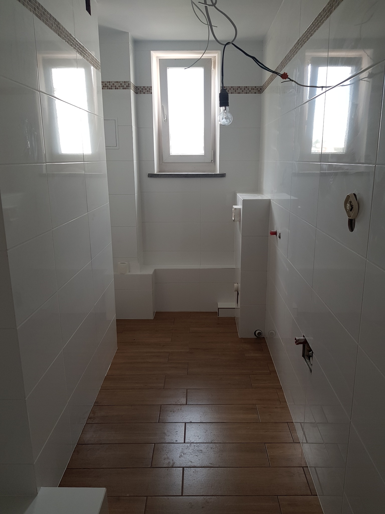 Wnętrze łazienki w trakcie prac wykończeniowych: białe płytki ścienne z dekoracyjnym pasem mozaiki, podłoga z płytek imitujących drewno, widoczne instalacje, surowa żarówka na kablu.