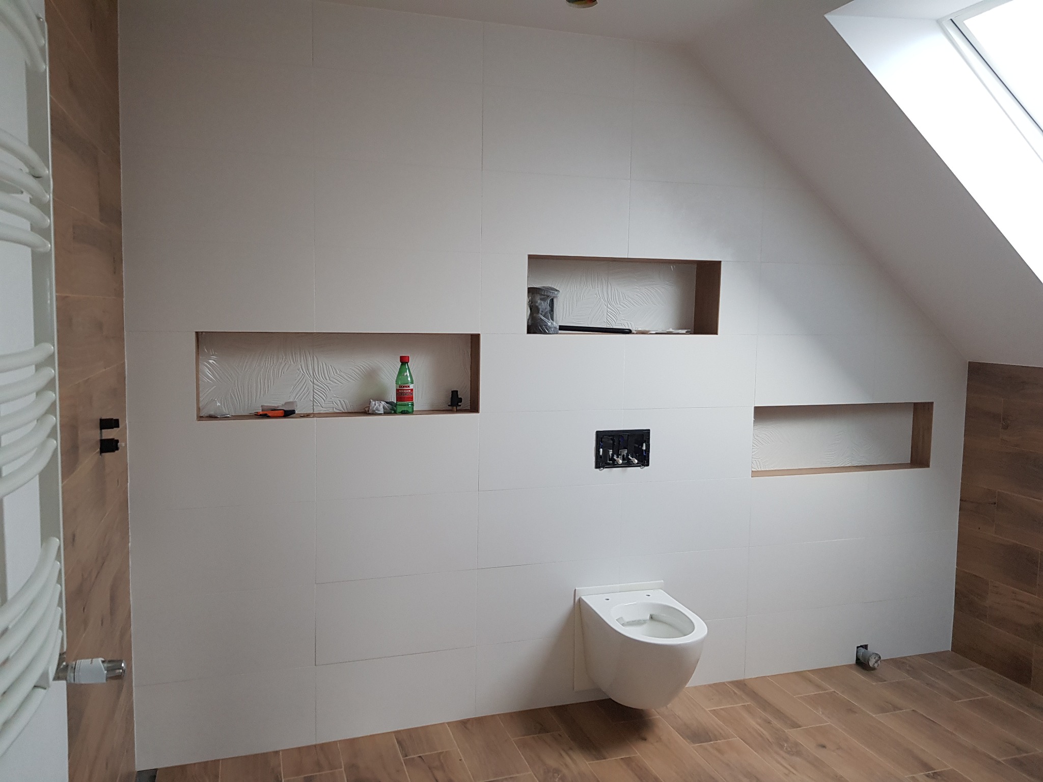 Białe płytki ścienne z wnękami w łazience na poddaszu, częściowo zamontowana toaleta wisząca, drewnopodobna podłoga, grzejnik drabinkowy po lewej stronie, butelka wody mineralnej w jednej z wnęk.