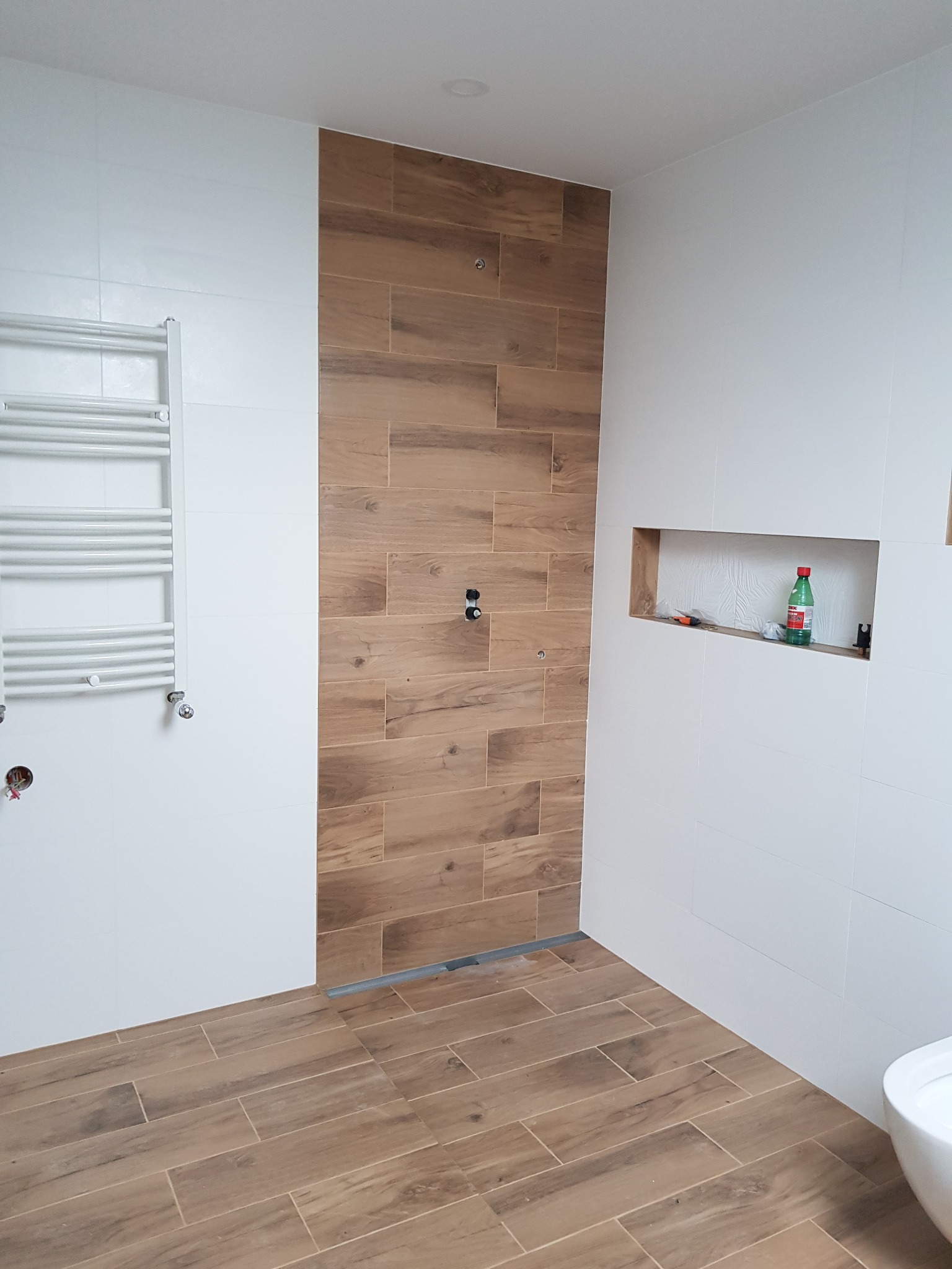 Łazienka w trakcie wykańczania: pionowy pas płytek imitujących drewno w strefie prysznica, białe płytki na pozostałych ścianach, wnęka w ścianie z butelką i narzędziami, grzejnik drabinkowy...