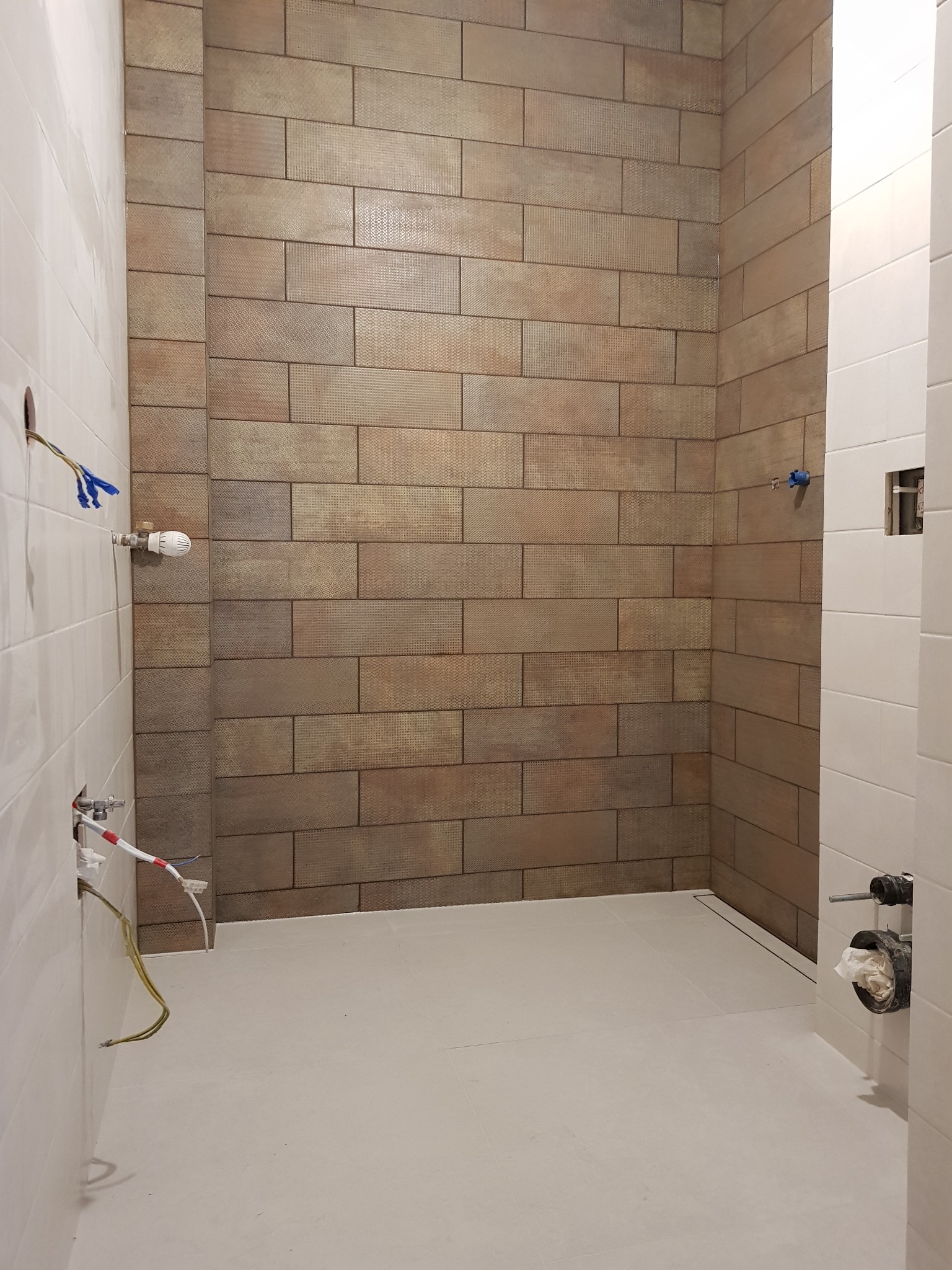 Ściany w trakcie układania płytek w łazience, widoczne przygotowane instalacje wodne i elektryczne, brązowe płytki imitujące cegłę na jednej ścianie, białe na drugiej, szary podkład pod posadzkę.