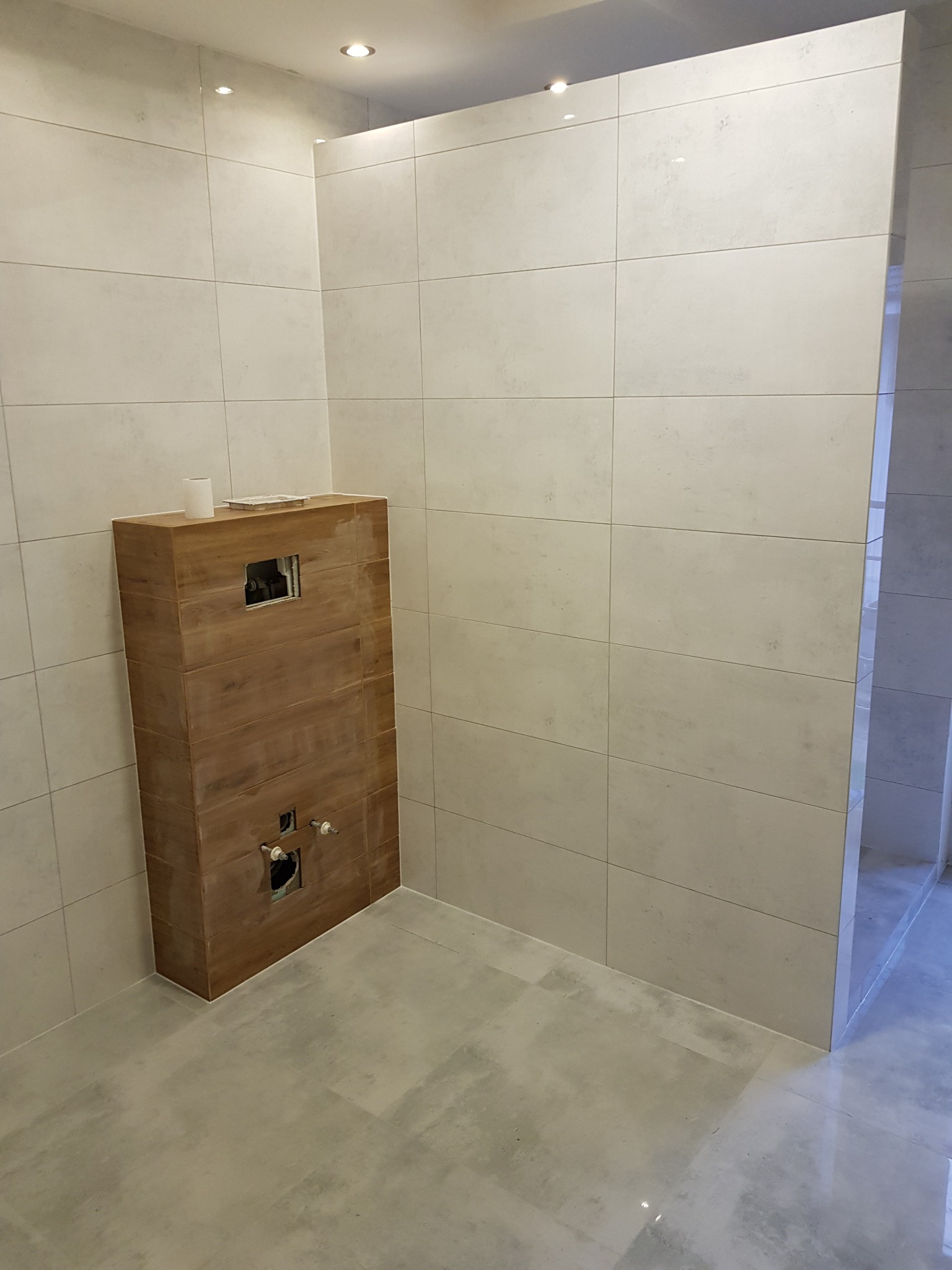 Niedokończona łazienka z drewnianą obudową stelaża podtynkowego WC, wyłożona jasnymi płytkami w formacie kwadratu.