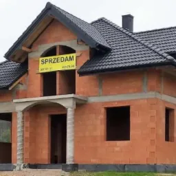 Niedokończony, dwukondygnacyjny dom z czerwonej cegły z czarnym dachem, widoczna tablica 'Sprzedam' na górnej kondygnacji i tablica informacyjna przed budynkiem.