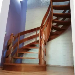 Kręcone schody z drewna o ciepłym odcieniu, z dekoracyjną spiralną poręczą, na tle ściany z białej cegły i fioletowej ściany.