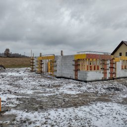 MATEUSZ ŚWIDER FIRMA BUDOWLANA - Wznoszenie ścian z bloczków betonowych na fundamencie domu jednorodzinnego, widoczne zbrojenie i szalunki, w tle ośnieżony teren i budynek mieszkalny.