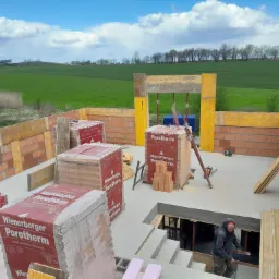 Budowa domu jednorodzinnego: Widok z góry na fundamenty z zalewanym betonem, częściowo w szalunkach, przygotowane palety z pustakami ceramicznymi Porotherm oraz pracownik schodzący po schodach...