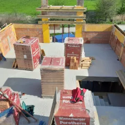 Wysoki kąt widzenia na plac budowy domu murowanego z widocznymi paletami cegieł Porotherm, szalunkami, podporami i otworem na schody w fundamencie.