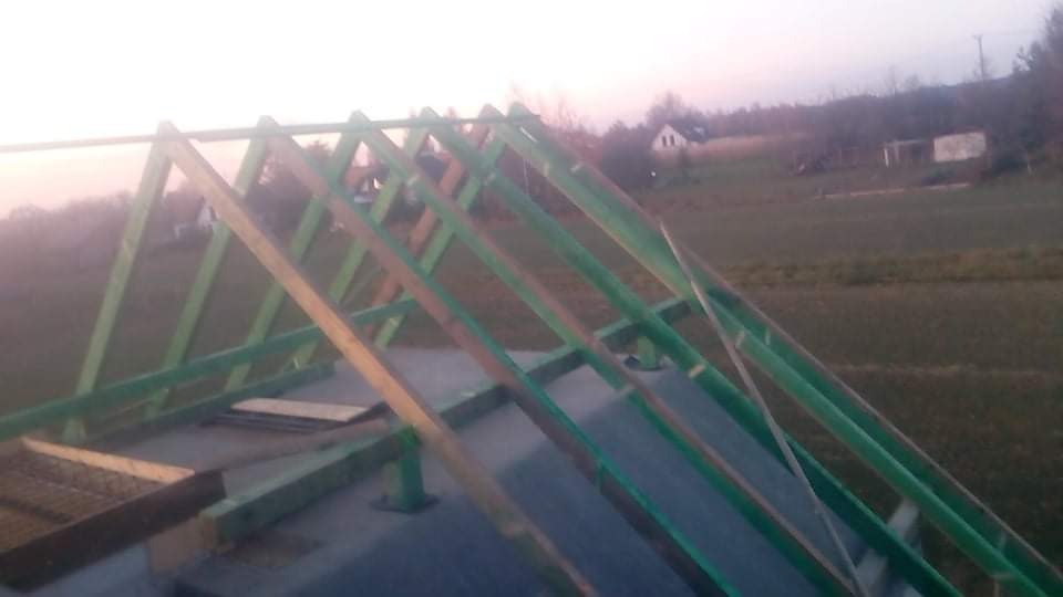 Konstrukcja drewniana dachu w trakcie budowy, z widocznymi elementami więźby pomalowanymi na zielono, na tle krajobrazu z domem i polami w oddali, uchwycona przy zachodzącym słońcu.