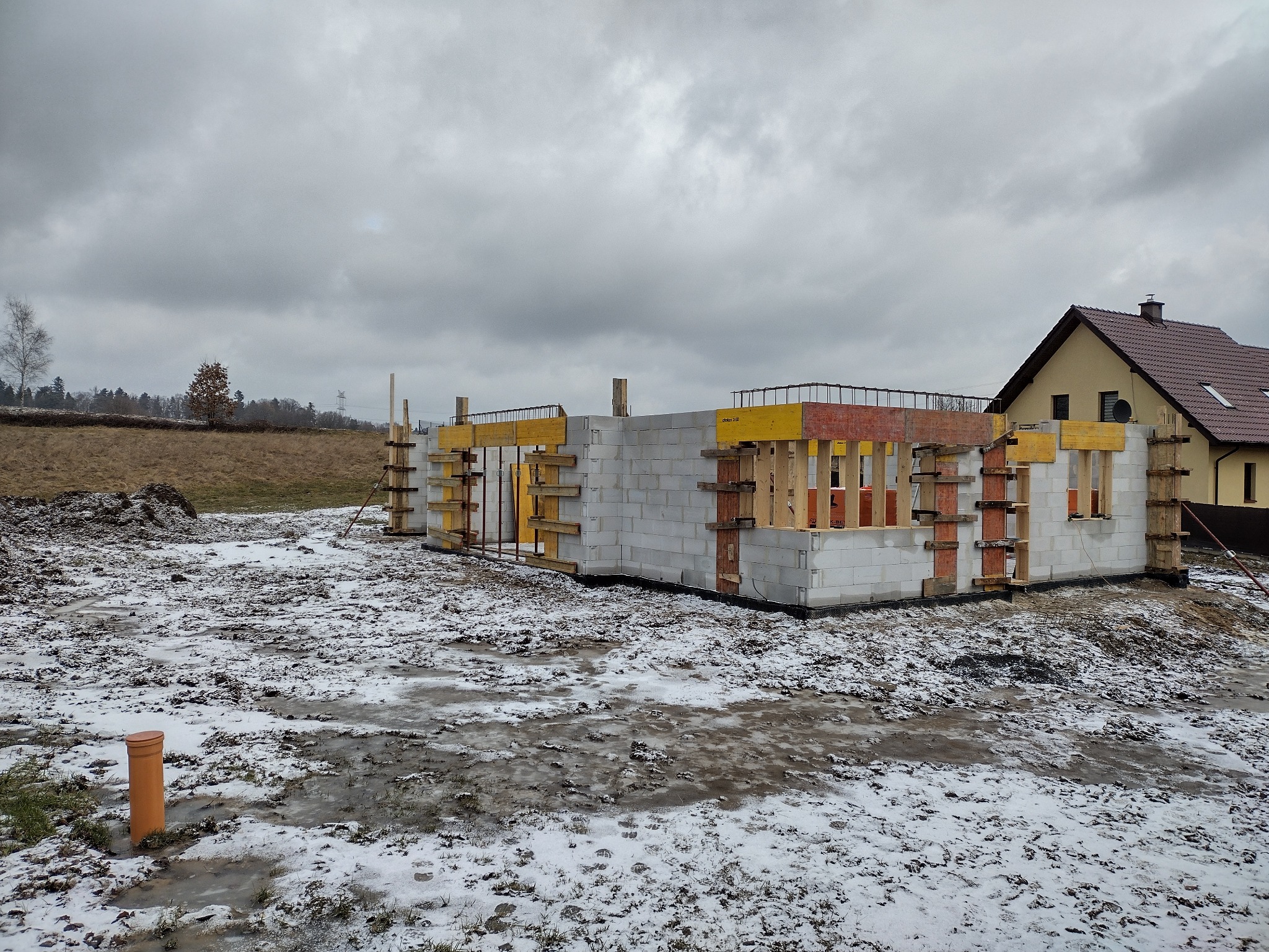 Wznoszenie ścian z bloczków betonowych na fundamencie domu jednorodzinnego, widoczne zbrojenie i szalunki, w tle ośnieżony teren i budynek mieszkalny.