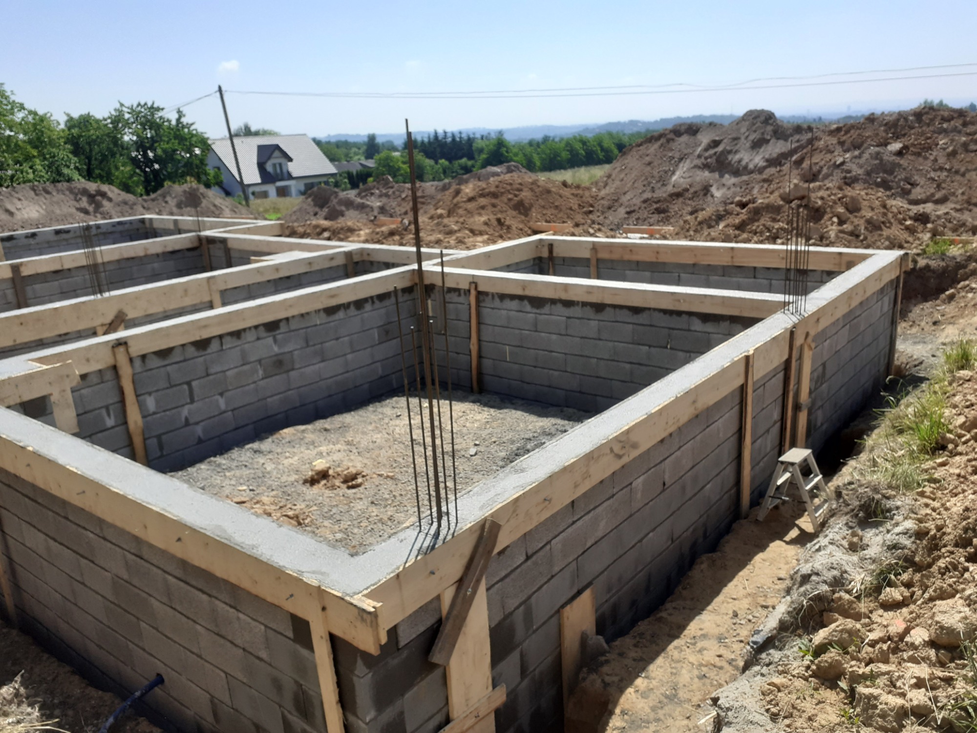 Wykopany i wylany fundament pod budynek z szarych bloczków betonowych, wzmocniony drewnianym szalunkiem i stalowymi prętami zbrojeniowymi, z widoczną stertą ziemi w tle i drabinką aluminiową z boku.