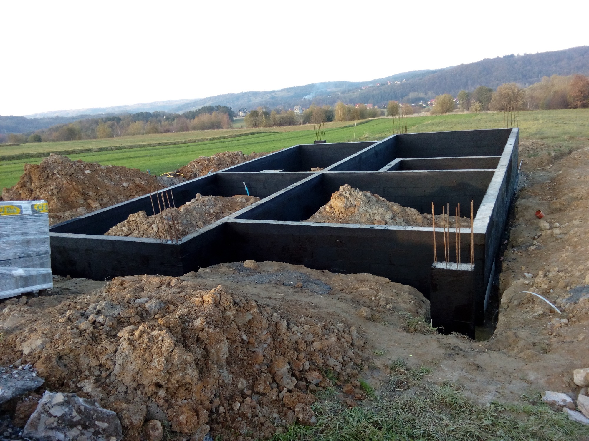Zabezpieczone hydroizolacją fundamenty domu jednorodzinnego w wykopie, z widocznymi prętami zbrojeniowymi i stertami ziemi na tle wiejskiego krajobrazu.