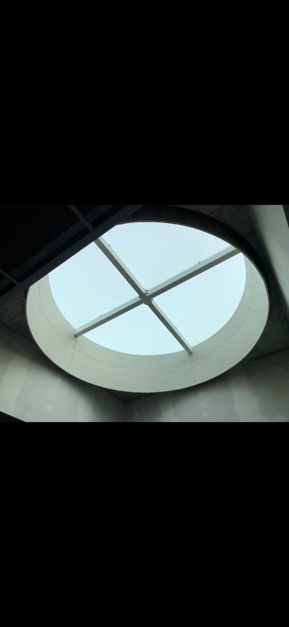 Owalne okno dachowe z białymi profilami krzyżowymi, widok od dołu, ściany w trakcie wykończenia.