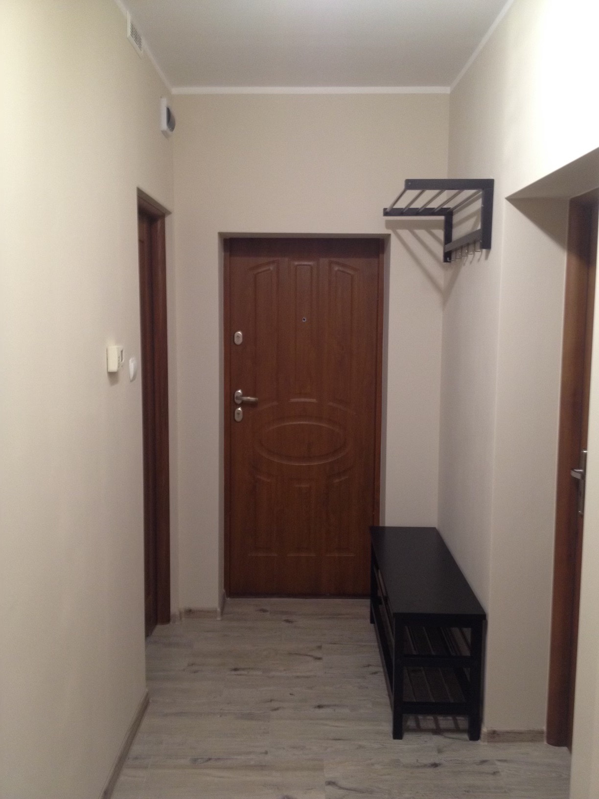 Przedpokój w mieszkaniu z brązowymi drzwiami wejściowymi, czarnym wieszakiem na ubrania i czarną ławką na buty, ściany pomalowane na jasny beż.