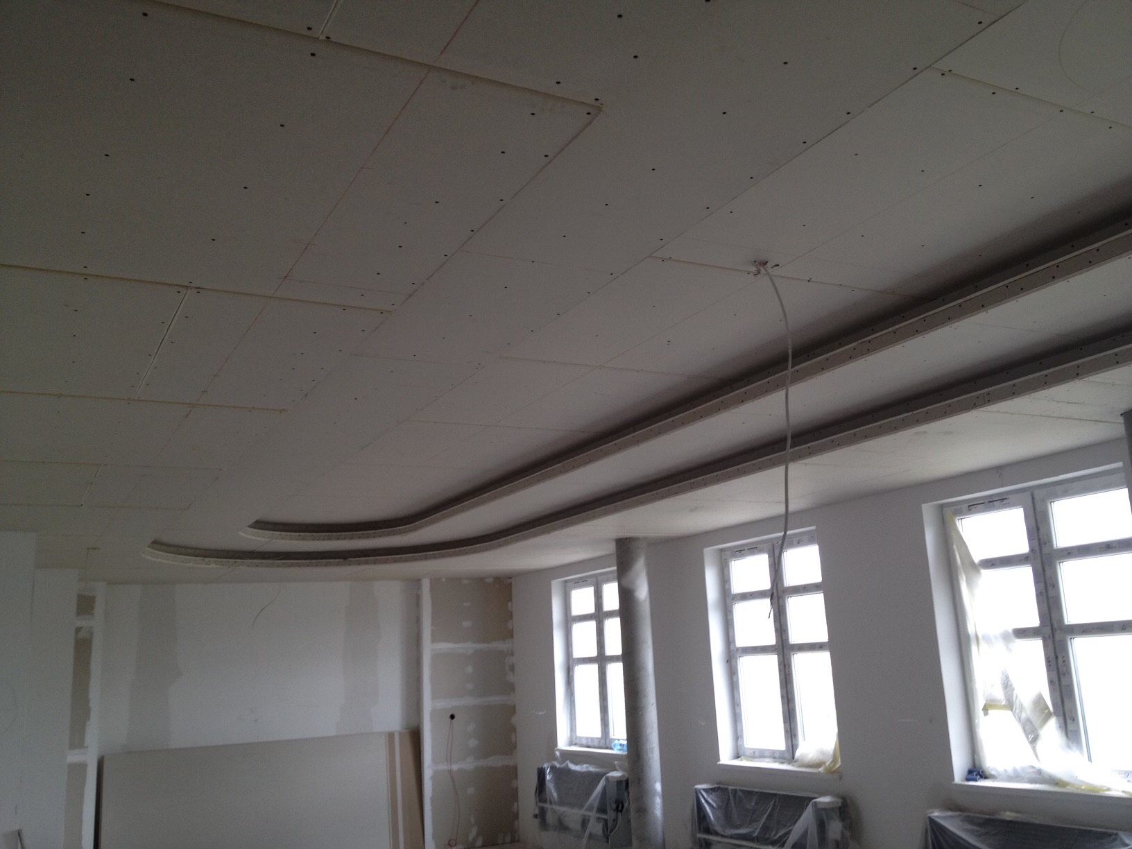 Sufit podwieszany z elementami dekoracyjnymi w trakcie montażu, widoczne profile i płyty, okna zabezpieczone folią, ściany w trakcie przygotowania do wykończenia.