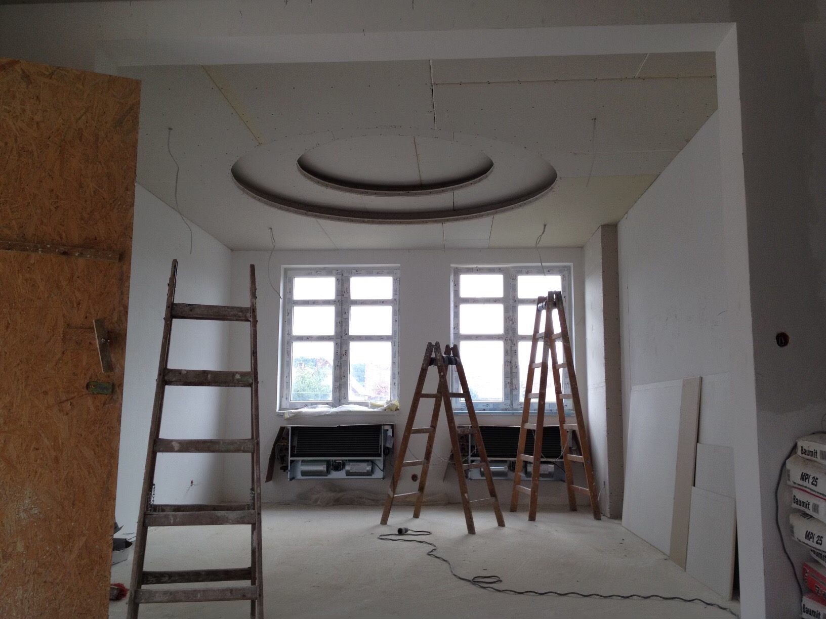 Wnętrze w trakcie prac wykończeniowych, widoczne drewniane drabiny, dwa okna, oraz sufit z okrągłym elementem dekoracyjnym i fragmentami niezabudowanymi.