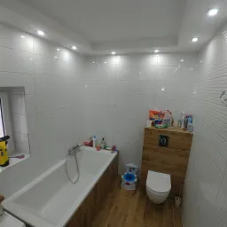 Nowoczesna łazienka z białymi, prostokątnymi płytkami na ścianach, drewnopodobnymi panelami na podłodze i obudowie toalety, oraz wanną z baterią.