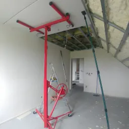 Czerwony podnośnik do płyt gipsowo-kartonowych podtrzymuje płytę przy suficie, widoczne profile sufitowe z izolacją, drabina i poziomica w tle, surowy stan wykończenia wnętrza.