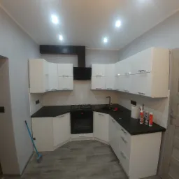 Nowoczesna kuchnia z białymi szafkami, czarnym blatem i drewnopodobną podłogą, widok ogólny z niebieską zmiotką opartą o ścianę.