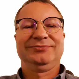 Portret uśmiechniętego mężczyzny w okularach z czarnymi oprawkami na białym tle, ubrany w szarą koszulę z kołnierzykiem.