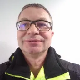 Portret hydraulika w okularach, ubrany w roboczą kurtkę z neonowo-zielonymi elementami, na tle białej ściany.