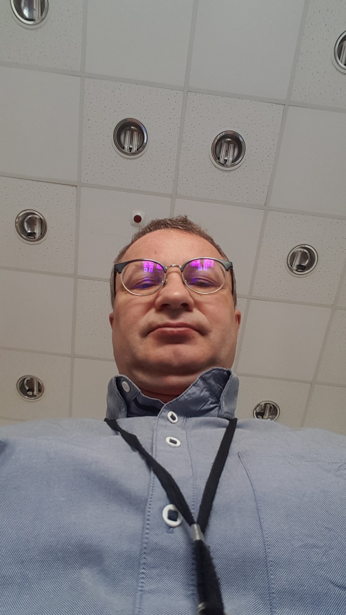 Portret mężczyzny w okularach, patrzącego z góry. W tle biały sufit kasetonowy z okrągłymi lampami. Mężczyzna ma na sobie błękitną koszulę z krótkim rękawem.