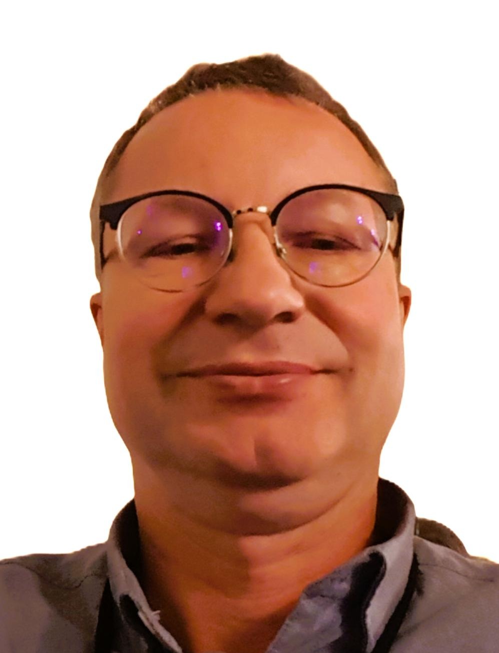 Portret uśmiechniętego mężczyzny w okularach z czarnymi oprawkami na białym tle, ubrany w szarą koszulę z kołnierzykiem.