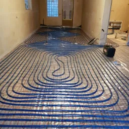 Niebieskie rury ogrzewania podłogowego ułożone na folii z siatką w surowym wnętrzu przed wylewką, widoczne okno z luksferami i grzejniki na ścianie w tle.