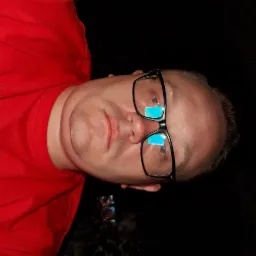 Portret mężczyzny w okularach z niebieskimi refleksami, ubrany w czerwoną koszulkę, na ciemnym tle.