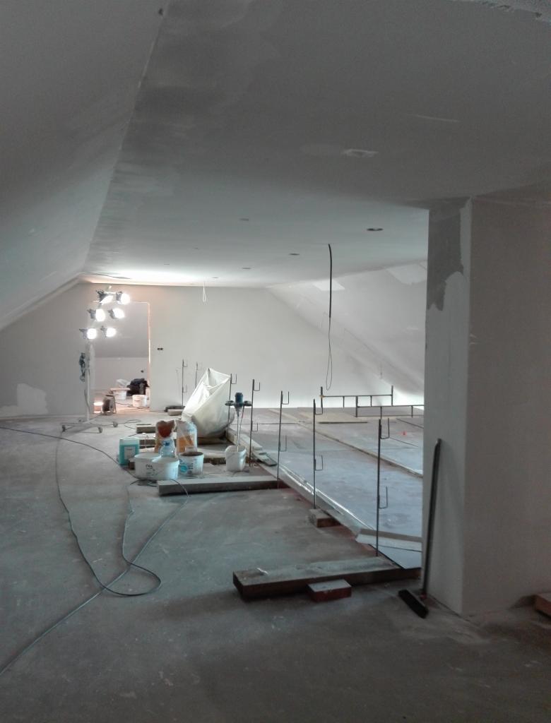 Poddasze w trakcie prac wykończeniowych: widoczne szpachlowane ściany i sufit, narzędzia, wiadra z farbą oraz konstrukcja pod przyszłą podłogę, oświetlenie budowlane.