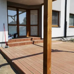 Nowoczesny taras z deski kompozytowej w kolorze brązowym, z niskimi schodami prowadzącymi do przeszklonego narożnika budynku, częściowo zadaszony drewnianą konstrukcją, widoczna szczotka...