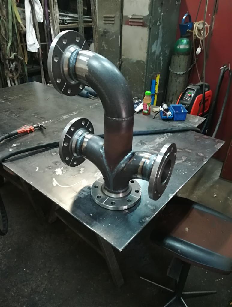 Zespawany trójnik z rur stalowych z kołnierzami, leżący na metalowym stole w warsztacie, widoczne ślady spawania i narzędzia.