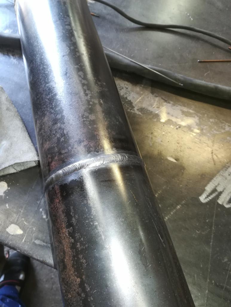 Połączenie spawane na rurze ze stali nierdzewnej, widoczne ślady po spawaniu i czyszczeniu, ułożone na metalowym stole warsztatowym.