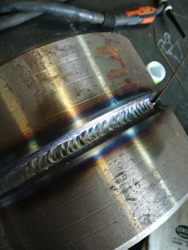 Metalowy cylinder z widocznym, świeżym spoinem z tęczowymi przebarwieniami, wykonanym metodą TIG, z elektrodą i uchwytem spawalniczym w tle.