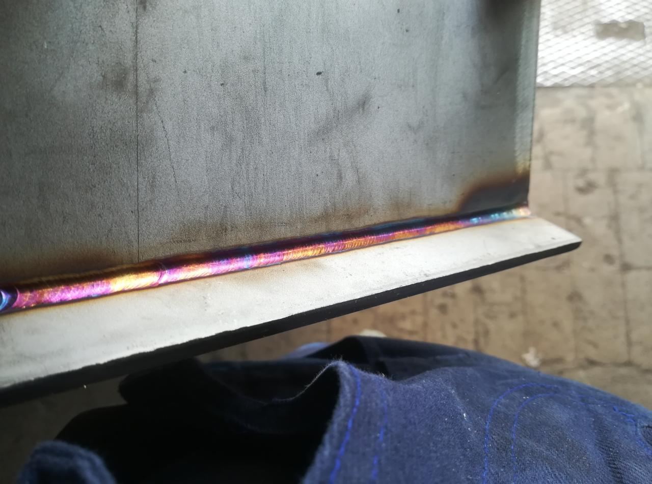 Metalowa konstrukcja z widocznym, opalizującym spoiwem po procesie łączenia, ułożona na ciemnoniebieskim materiale. Widoczne ślady obróbki na metalu.